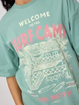 Remera blanca de algodón con estampado frontal de estilo playero con texto "Welcome to the Surf Camp" y dibujo de una casa de playa con palmeras y tablas de surf.