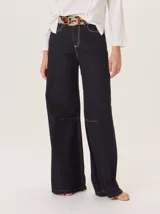 Pantalón ancho de denim oscuro, corte holgado, con pespuntes a contratono color naranja. Presenta detalles de pinzas por debajo de la rodilla y bordado de monograma en el bolsillo delantero.