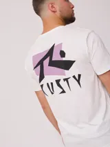 Remera blanca de algodón con estampado gráfico en la espalda. El diseño presenta el logo de Rusty en negro sobre un fondo rectangular lila, con tipografía angular.