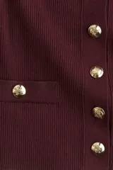 Cárdigan corto de punto acanalado en color vino tinto, con cuello redondo, manga corta y cierre frontal de botones dorados. Presenta dos bolsillos delanteros decorativos con botón dorado a la altura de la cadera.