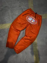 Conjunto puffer naranja con estampado del logo de la marca en negro. Incluye campera sin mangas con mangas desmontables, pantalón de jogging, gorro y tote bag. Resistente al agua y al frío.
