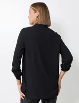 Blusa negra de manga larga con escote en V y pliegue central. La tela presenta un tejido piqué texturado sutil. Las mangas se pueden remangar y sujetar con una presilla y botón dorado.