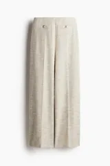 Pantalón de corte holgado color beige, en tejido texturizado con pliegues adelante y atrás. Modelo de tiro alto con elástico oculto en la cintura y piernas anchas. Bolsillos delanteros decorativos con botón de metal con diseño.