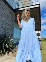 Vestido camisero largo celeste con rayas verticales blancas, cuello camisero, manga larga con puño y abotonadura frontal. Diseño escalonado con volantes en cada corte.