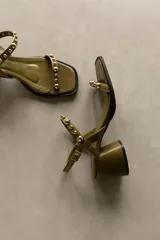 Sandalia de cuero color marrón con taco bloque medio y punta cuadrada. Presenta tachas esféricas doradas en las tiras y hebilla de ajuste en el tobillo.