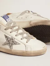 Championes Golden Goose modelo Super-Star confeccionados en cuero blanco con efecto desgastado. Destacan por su estrella lateral en glitter plateado, refuerzo del talón en cuero marrón y forro interior de lana tipo shearling. Incluyen cordones grises y suela de goma con acabado envejecido.