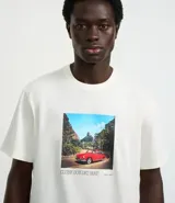 Remera de algodón color crema con cuello redondo y mangas cortas. Presenta un estampado rectangular con la imagen de un auto rojo en un paisaje y texto en la parte inferior.