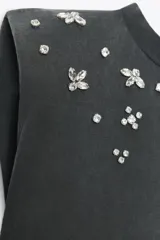 Camiseta negra con efecto lavado, cuello redondo, sisas amplias con hombreras y apliques de joyas combinadas a contraste en la parte superior.