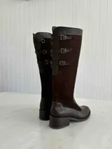 Bota alta de cuero negro con caña alta y taco cuadrado. Presenta tres correas con hebillas plateadas en la parte superior de la caña. Fabricada en Colombia.