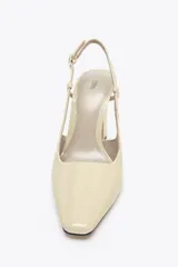 Zapato de tacón destalonado color beige acharolado, con punta fina, tacón ancho en bloque y cierre mediante tira trasera con hebilla ajustable.