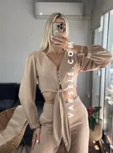 Conjunto color beige compuesto por un top corto con escote en V, botones frontales y mangas largas abullonadas con puños elásticos, y un pantalón de tiro alto con lazo en la cintura.