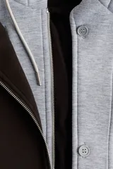 Campera negra de tejido suave efecto neopreno, con cuello y cierre frontal. Presenta un diseño de doble capa simulado con una capucha de tela gris claro que asoma por debajo del cuello de la campera.