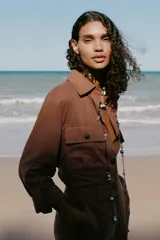 Camisa color marrón con cuello solapa y manga larga acabada en vuelta con trabilla y botón. Bolsillos delanteros de plastrón con solapa. Bajo con aberturas laterales. Cierre frontal con botones y cinturón de la misma tela.