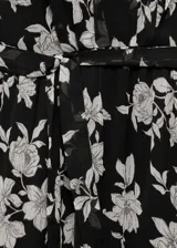 Vestido corto negro con estampado floral blanco, de mangas largas acampanadas, cuello redondo, lazo en la cintura y volados en el ruedo.
