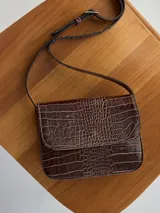 Mini cartera marrón con textura de cocodrilo, correa larga regulable y cierre con solapa.