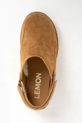 Zueco tipo mule de gamuza color camel, con forro interior de corderito visible en el borde. Presenta una tira ajustable con broche metálico en el lateral y suela de goma gruesa y dentada.