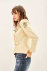 Saco sastre color beige claro, de manga larga y calce al cuerpo, con cierre frontal ajustable mediante cinturón con hebilla de piedras. Presenta bordados florales en tonos dorados y negros con lentejuelas y canutillos en los hombros y solapas.