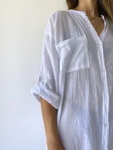 Vestido camisero blanco de corte holgado, con cuello mao, cierre frontal con botones, mangas 3/4 con puños abotonados, dos bolsillos de parche en el pecho y aberturas laterales en el ruedo.