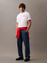 Remera blanca de algodón con cuello redondo, mangas cortas y logo de Calvin Klein Jeans estampado verticalmente en color rojo en el pecho.