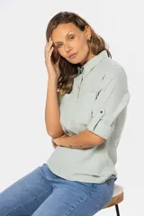 Camisa color beige de lino, de manga larga con botones metálicos personalizados y presilla en la manga. Tiene cuello clásico, cierre frontal parcial con botones y bolsillo de parche en el pecho.