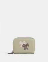Billetera pequeña de cuerina color blanco con estampado de lazos rosas y corazones. Tiene cierre circundante y tirador de goma.