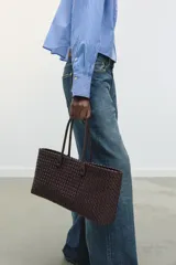 Bolso shopper alargado color marrón chocolate con diseño trenzado. Tiene doble asa de hombro y cierre superior con botón imantado. Incluye una bolsa interior con cierre fruncido.