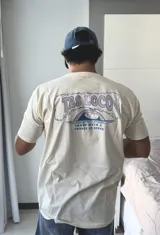 Remera de manga corta color beige jaspeado, con un estampado gráfico en la espalda que incluye el texto "TAS LOCO" y "SUNNY WITH A CHANCE OF STOKE" con un diseño de una persona en el agua con una ballena.