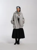 Buzo hoodie oversize de felpa de algodón color gris, con capucha y bufanda integrada que envuelve el cuello. Presenta cierre frontal y bolsillos canguro.