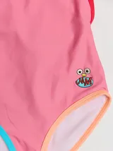 Traje de baño entero rojo con tirantes finos y espalda cruzada. Presenta un parche bordado con un monstruo en la parte inferior. Tiene ribetes en color borgoña y celeste en la parte superior, y ribetes en color naranja y verde en la parte inferior.