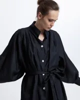 Vestido camisero negro de ramio con cuello mao, mangas largas abullonadas, cierre frontal con botones blancos y lazo a la cintura.