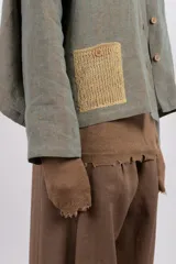 Camisa amplia de lino en tono verde desgastado, con textura visual y caída liviana. Destaca por su parche tejido a mano en contraste, que aporta un detalle artesanal y único.