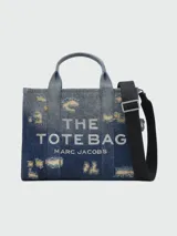 Bolso tote Marc Jacobs de tamaño mediano confeccionado en denim azul con efecto desgastado. Presenta el logo "THE TOTE BAG MARC JACOBS" estampado en blanco en el frente, doble asa corta superior y correa bandolera extraíble de tela negra.