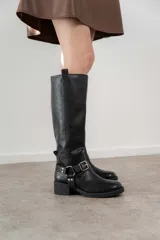 Bota de caña alta color negro, con hebilla y arnés decorativo en el tobillo, y taco bajo. Confeccionada en material sintético PU (poliuretano), libre de componentes de origen animal.