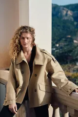 Gabardina corta color beige con doble botonadura, cuello clásico, charreteras en los hombros, bolsillos delanteros con solapa, mangas anchas con trabillas ajustables y pliegue en la espalda.