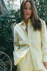 Camisa tipo pijamera a rayas amarillas y blancas, confeccionada en hilatura con algodón y lino. Tiene cuello solapa, manga larga con puños y cierre frontal con botones.