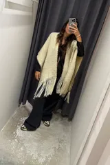 Ruana o poncho de tejido grueso color beige claro, con flecos largos en el borde inferior y ribete de costura en contraste en la abertura frontal.