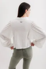 Camisa blanca de viscosa con cuello camisero, mangas largas abullonadas con puños elásticos y cierre frontal con botones. Presenta bordados negros con motivos florales en las mangas.