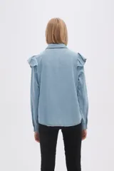Camisa de jean azul con cuello clásico, cierre frontal con botones y mangas largas. Presenta volados en el canesú delantero y trasero.