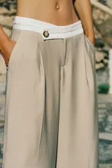 Pantalón color beige de tiro alto con cintura combinada en blanco, pinzas delanteras y pernera ancha. Cierre frontal con cremallera y botón.