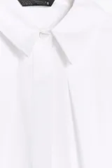 Camisa blanca de popelín con cuello solapa y sisa. Presenta pinzas delanteras y cierre frontal con botones ocultos por solapa.