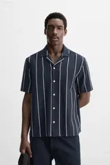 Camisa de manga corta con cuello bowling y corte relaxed fit. Diseño a rayas verticales en tonos beige y blanco. Cierre frontal con botones y bajo con aberturas laterales.