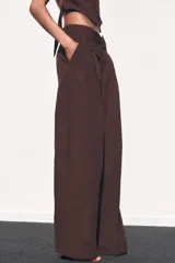 Pantalón marrón de tiro medio con trabillas, bolsillos laterales y de vivo en espalda. Detalle de pliegues y cierre frontal con cremallera, botón interior y gancho metálico.