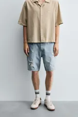 Bermuda de denim de algodón, color gris lavado, con corte relaxed fit y roturas visibles en la parte delantera de ambas piernas. Presenta cinco bolsillos y cierre frontal con cremallera y botón.