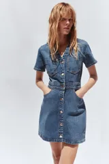 Vestido corto de denim azul, con cuello solapa, escote en pico, manga corta, bolsillos delanteros y cierre frontal con botones metálicos.