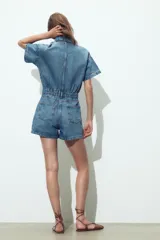 Mono corto de denim azul, con cuello solapa, manga corta, bolsillos delanteros y traseros, cintura elástica en la espalda y cierre frontal con cremallera y botones metálicos.