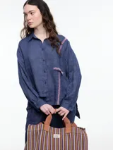 Camisa de manga larga de lino color azul con calce relajado y diseño asimétrico. El bolsillo delantero está colocado de forma invertida, un detalle lúdico que rompe con lo clásico. Lleva un bordado tipo pespunte en color contraste que recorre el frente y el bolsillo.