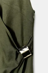 Vestido midi color verde oliva confeccionado en lino. Presenta escote pico cruzado, tirantes anchos delanteros y tirantes finos en la espalda, detalle de aplique dorado en la cintura, bajo con abertura frontal y cierre lateral con cremallera oculta.