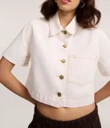 Camisa cropped de sarga color crudo, con cuello clásico, cierre frontal con botones dorados, manga corta, bolsillo de parche en el pecho y costuras a contraste. Confeccionada con algodón certificado.