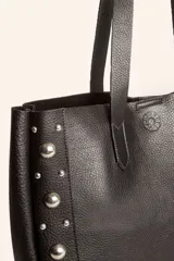 Bolso tote de cuero color marrón con tachas plateadas en los laterales. Tiene doble asa de hombro y un sobre interior de cuero.