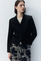 Blazer negro de punto pesado con solapas de muesca, corte entallado, hombreras, bolsillos delanteros ribeteados y abertura decorativa en los puños.